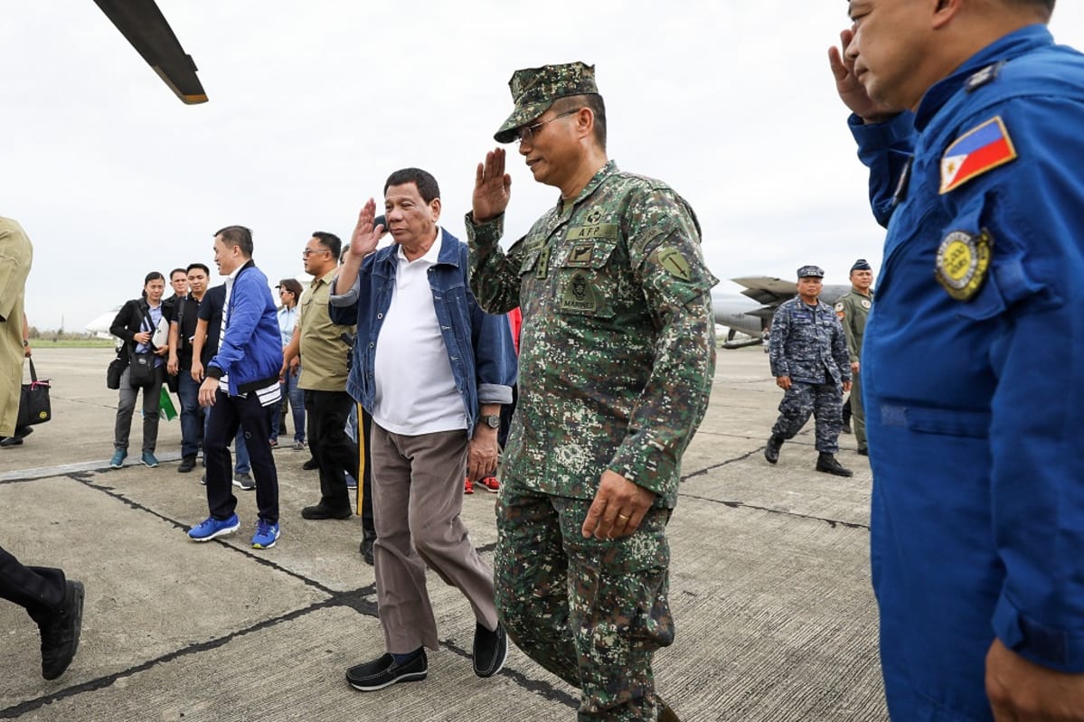 Rodrigo Duterte kävelee lentokentällä valkoisessa paidassa ja farkkutakissa. Presidentti ja hänen vieressään kävelevä sotilashenkilö tekevät juuri kunniaa. Ympärillä näkyy sotilaita, toimittajia, valokuvaajia. Kuvan ylälaidassa näkyy helikopterin lapa.