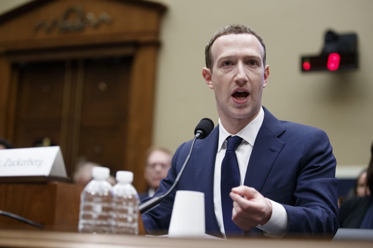 Tummansiniseen pukuun, valkoiseen kauluspaitaan ja tummansiniseen kravattiin pukeutunut Zuckerberg istuu pöydän takana ja puhuu mikrofoniin. Taustalla näkyy iso ovi. Pöydällä on kaksi vesipulloa.
