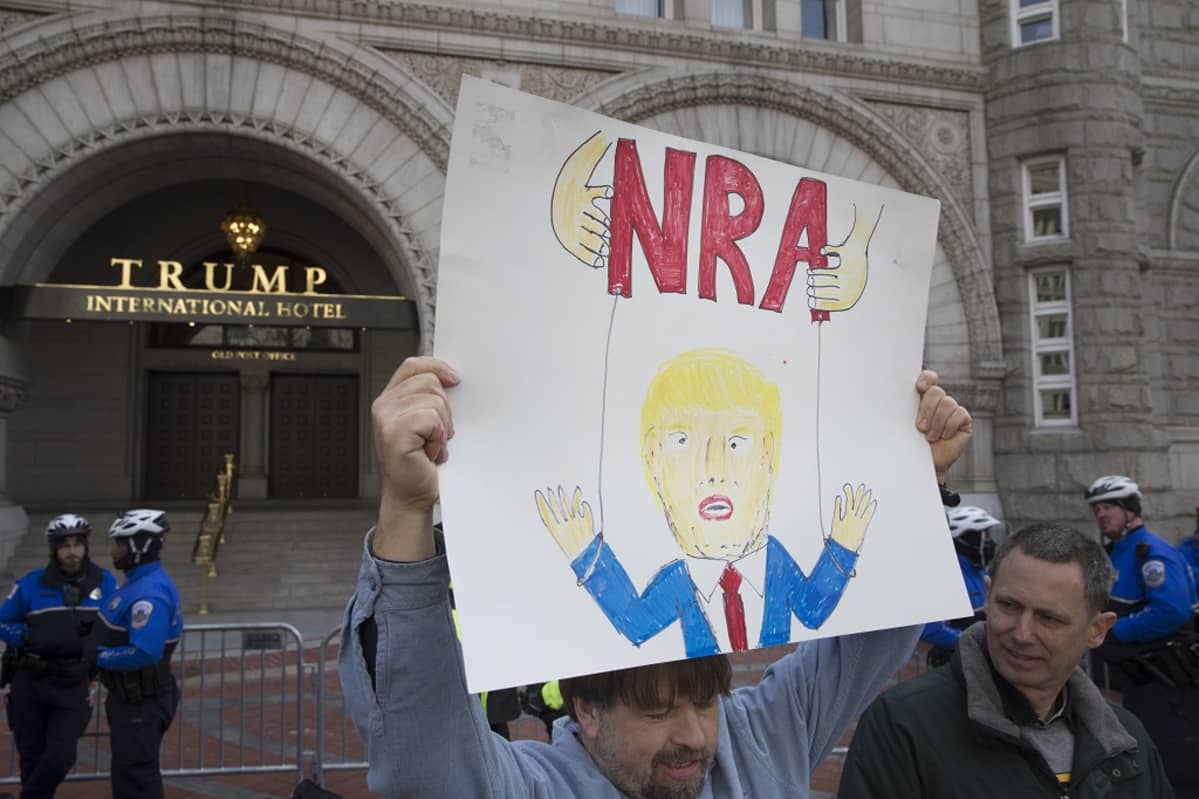 Kuvassa mielenosoittaja kannattelee kylttiä, jossa on Donald Trumpin kuva ja teksti NRA. Viesti on, että NRA kontrolloi Trumpia.