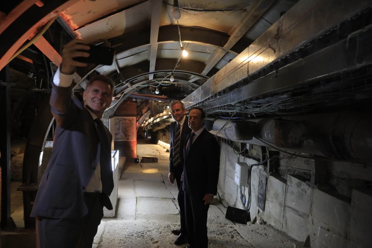 Kolme miestä tunnelissa, etummainen ottaa kännykällä selfietä.