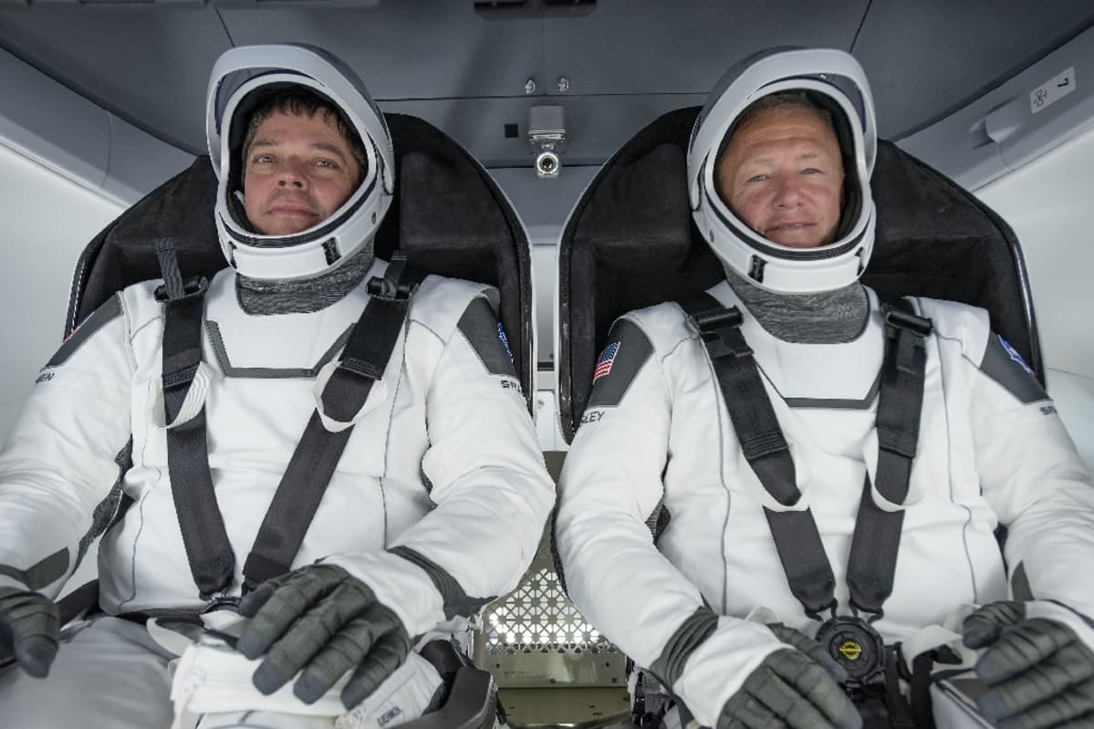 Kaksi valkopukuista astronauttia turvavyöt kiinnitettyinä istuimissaan avaruuskapselissa.