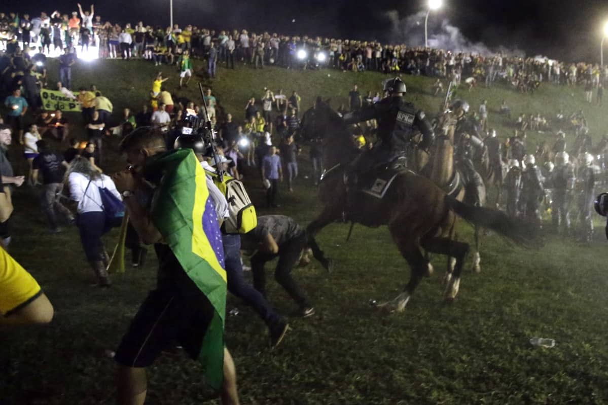 Iltaisessa tai yöllisessä kuvassa ratsupoliisi karauttaa hevosellaan mielenosoittajien joukkoon. Ihmiset juoksevat poispäin poliiisista. Etualalla on Brasilian lippuun kääriytynyt mielenosoittaja. Taustalla harjanteen päällä näkyy mielenosoittajien rivistöä. Joukko muita mellakkapoliiseja on harjanteen alaosassa. Ilmassa leijuu kyynelkaasua.