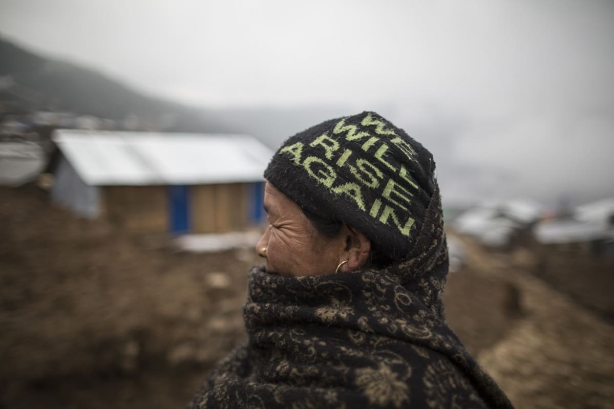 Iäkäs nainen katselee maisemaa. Hänellä on kaulansa ja leukansa peittävä paksu tumma huivi ja tumma pipo, jossa lukee vihreillä kirjaimilla 'WE WILL RISE AGAIN'. Taustalla näkyy hökkeleitä sumuisessa vuoristomaisemassa.