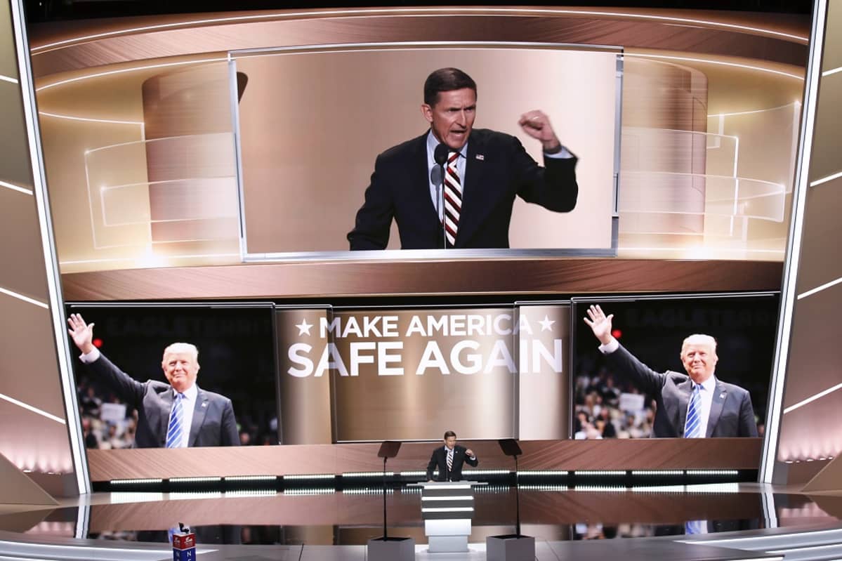Michael Flynn puhuu korokkeen takaa puoluekokousareenalla. Kuvassa hän näkyy pienenä hahmona ja valtavana kuvana takanaan olevalla suurella näytöllä. Näytön alareunassa on kaksi kuvaa Trumpista ja niiden välissä teksti MAKE AMERICA SAFE AGAIN. Flynn pitää vasenta nyrkkiään pystyssä. Hänellä on musta puju, raidallinen kravatti ja Yhdysvaltain lippua esittävä pinssi takkinsa kauluksessa. Hänen ilmeensä on vihainen.