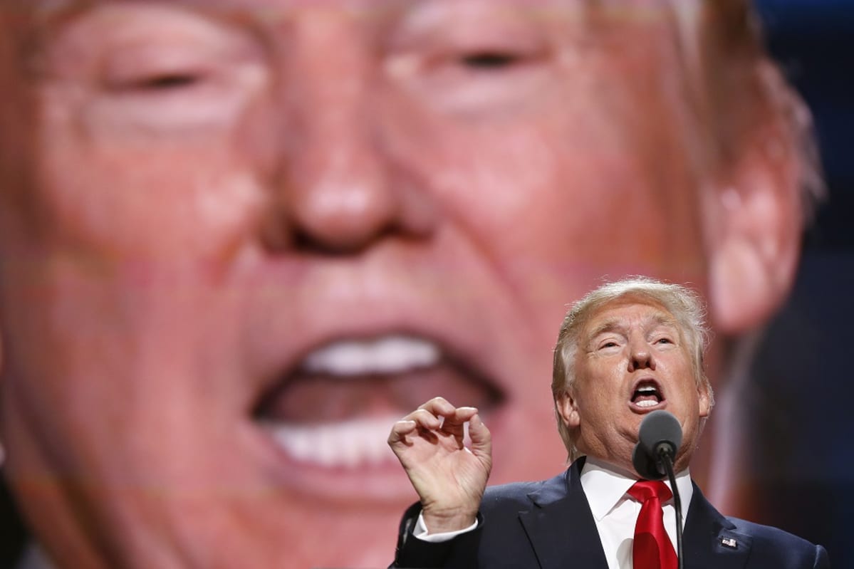 Trump puhuu valtavan näytön edessä mikrofoniin. Hän tehostaa sanojaan oikealla kädellään, etusormi ja peukalo yhdessä. Hänellä on tumma puku, kirkkaanpunainen kravatti. Taustalla näyttö esittää Trumpin puhetta. Trumpin kasvokuva täyttää koko taustan.