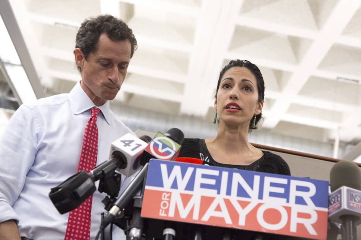 Huma Abedin puhuu mikrofoneille. Vieressä Anthony Weiner puree huultaan.