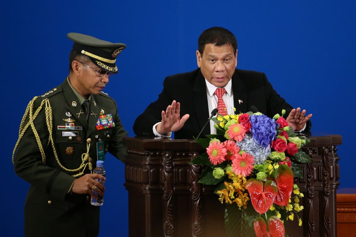 Rodrigo Duterte seisoo kukilla koristellun puhujanpöntön takana ja puhuu mikrofoniin molemmat kädet hieman kohotettuina. Presidentillä on musta puku ja punainen valkopilkkuinen kravatti. Vieressä sotilas tuo muovista vesipulloa. Tausta on syvänsininen.