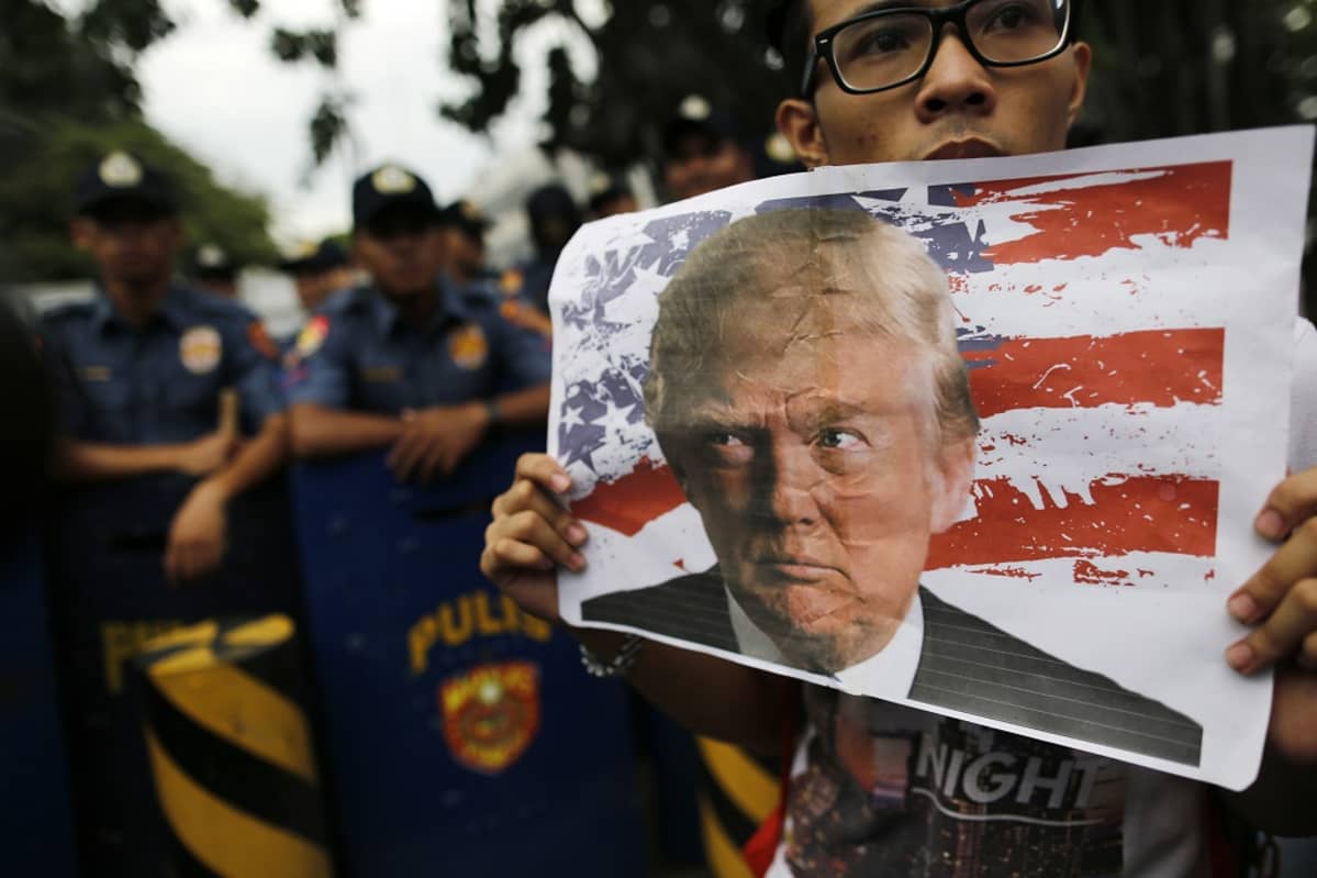 Silmälasipäinen mielenosoittaja pitelee Donald Trumpin kuvaa. Taustalla näkyy poliiseja tarkkailemassa tilannetta.