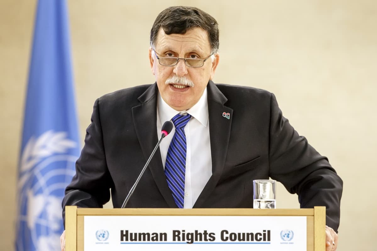 Sarraj seisoo puhujanpöntön takana ja puhuu mikrofoniin. Hänellä on tumma puku ja siniraitainen kravatti. Sarrajilla on harmaat viikset, tumma tukka ja silmälasit. Taustalla näkyy YK:n sininen lippu.