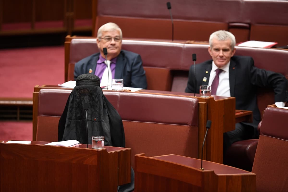 Kuvassa Pauline Hanson istuu parlamentissa burga päällään. Hänen takanaan istuu kaksi miestä.
