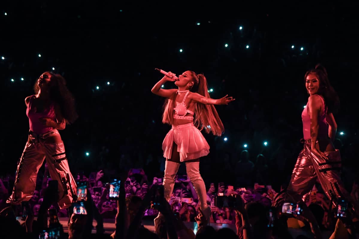 Laulaja Ariana Grande laulaa silmät kiinni lavalla kahden tanssijan ympäröimänä. Hänellä on yllään valkoinen latex-toppi, lyhyt vaalea röyhelöminihame ja korkeat valkoiset latex-saappaat.