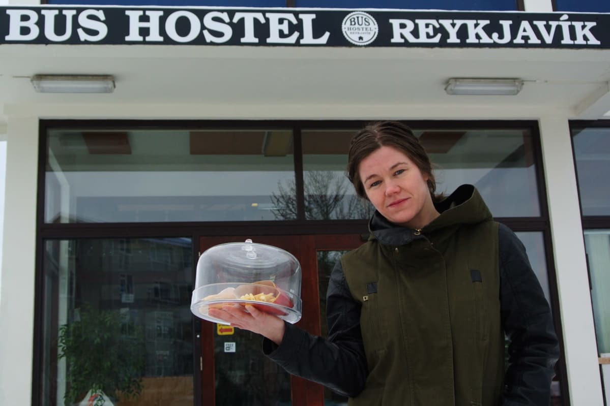 Nainen näyttää lasikuvun alla olevaa hampurilaista. Taustalla hostellin nimikyltti.