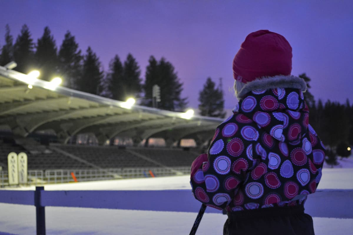 Lapsi seisoo hiihtosauvojen kanssa ja katselee urheilustadionin valoja.