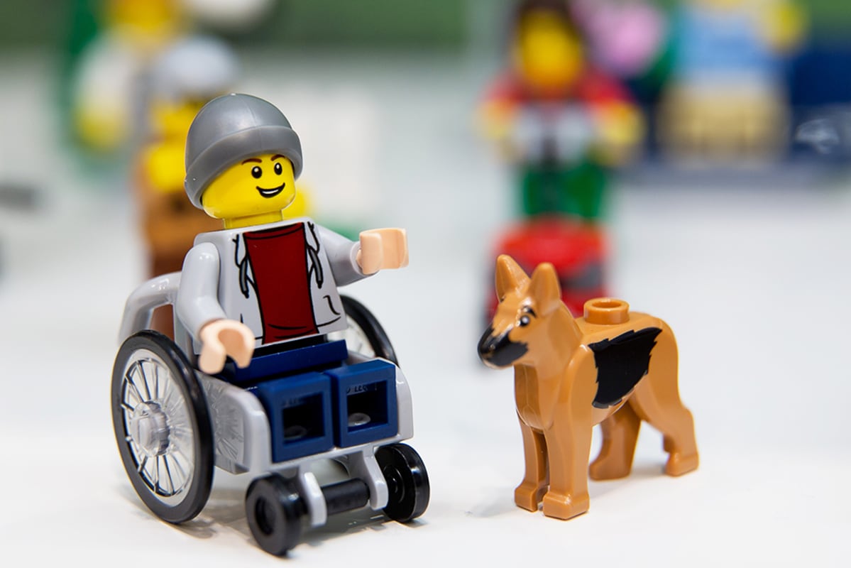 Pyörätuolissa istuva Lego-hahmo avustajakoiran seurassa.