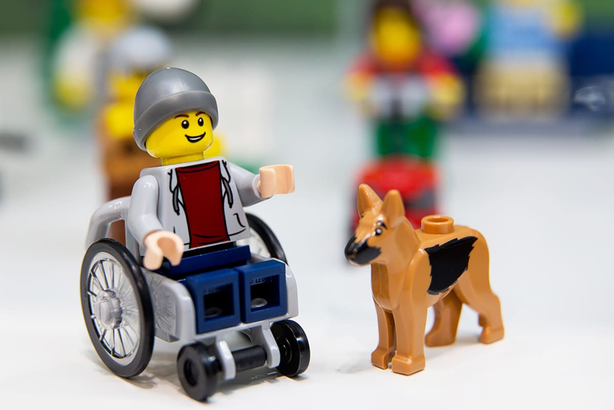 Pyörätuolissa istuva Lego-hahmo avustajakoiran seurassa.