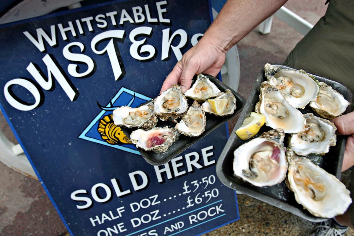 Miehellä on kädessään kaksi lautasannosta ostereita. Takana Whitstable oysters -mainos.