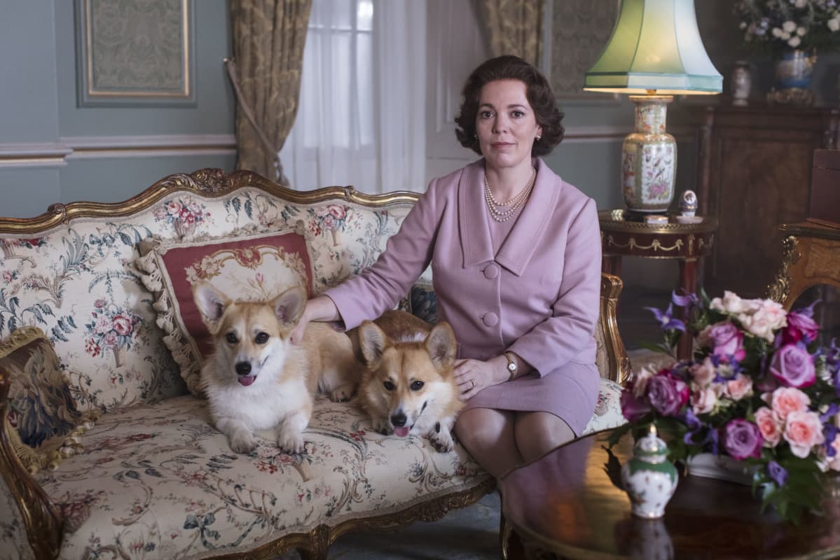 Olivia Colman som spelar drottning Elizabeth sitter i en antik soffa i en vacker salong med två av sina Corgis intill sig.