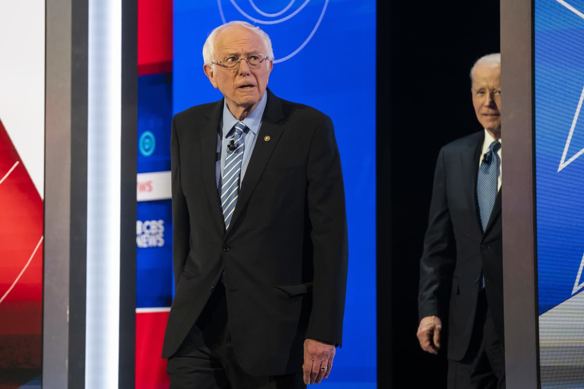Bernie Sanders kävelee lavalle, perässä Joe Biden oviaukossa.