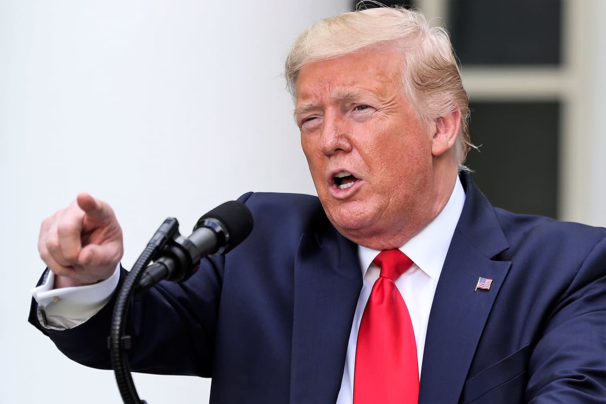 Donald Trump puhuu edessään olevaan mikrofoniin ja osoittaa etusormellaan yleisön suuntaan. Hänellä on tumma puku ja punainen kravatti.