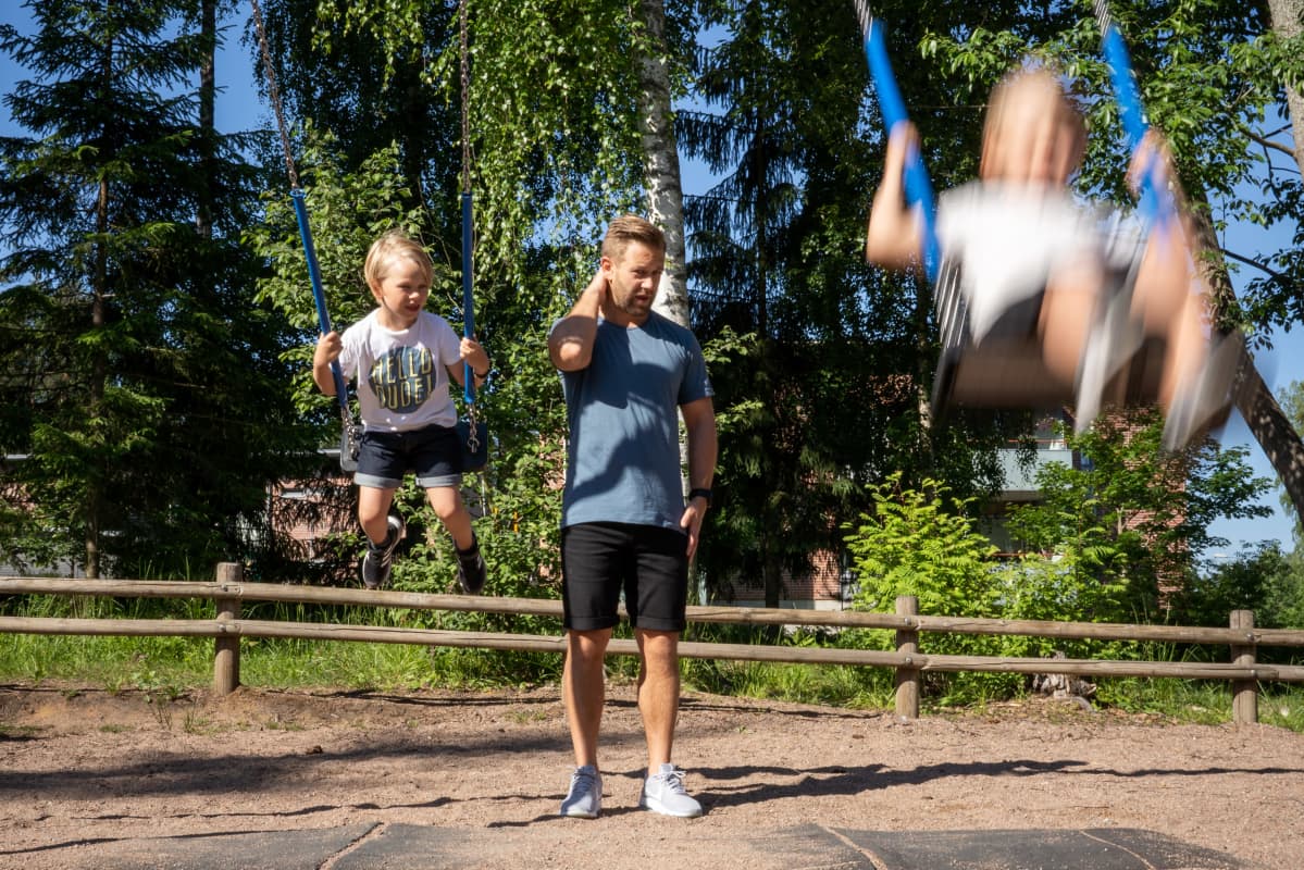 Mies seisoo keinujen takana ja hieroo niskaansa. Molemmin puolin keinuvat pojat kovaa vauhtia.