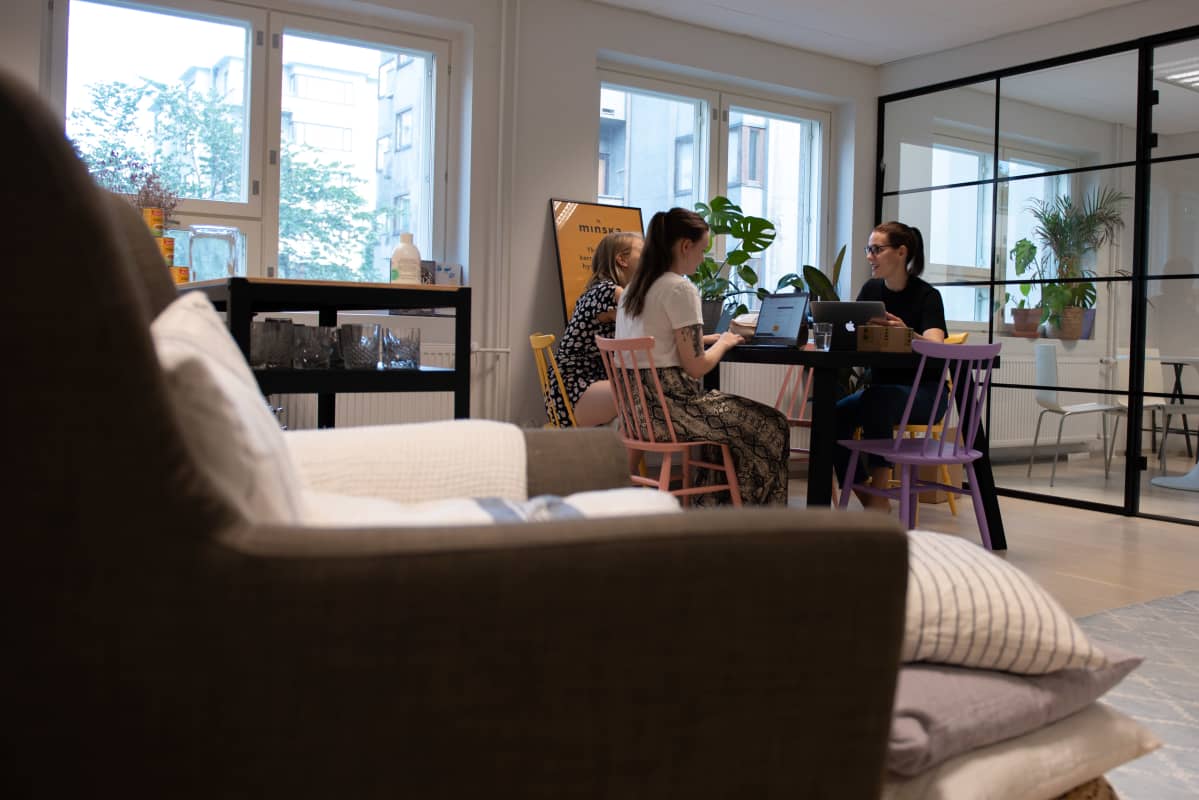 Outi Ellilä, Annika Ikonen ja Hetta Ahokas pitävät kokousta pöydän ääressä ikkunan edessä. Etualla näkyy nojatuoli.