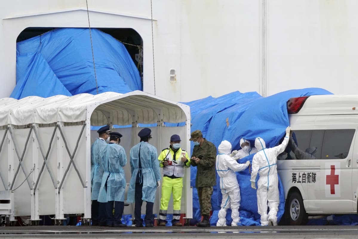 Suojapukuihin pukeutuneet ihmiset vetävät laivan kyljessä olevasta aukosta tulevaa sinistä suojakangasta ambulanssin päälle.