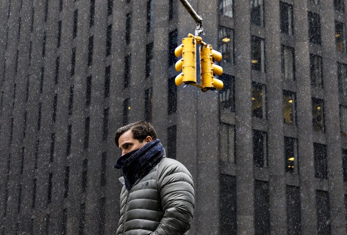 Mies kävelee lumisateessa urbaanissa New Yorkissa, jonka harmauden rikkovat keltaiseksi maalatut liikennevalot.