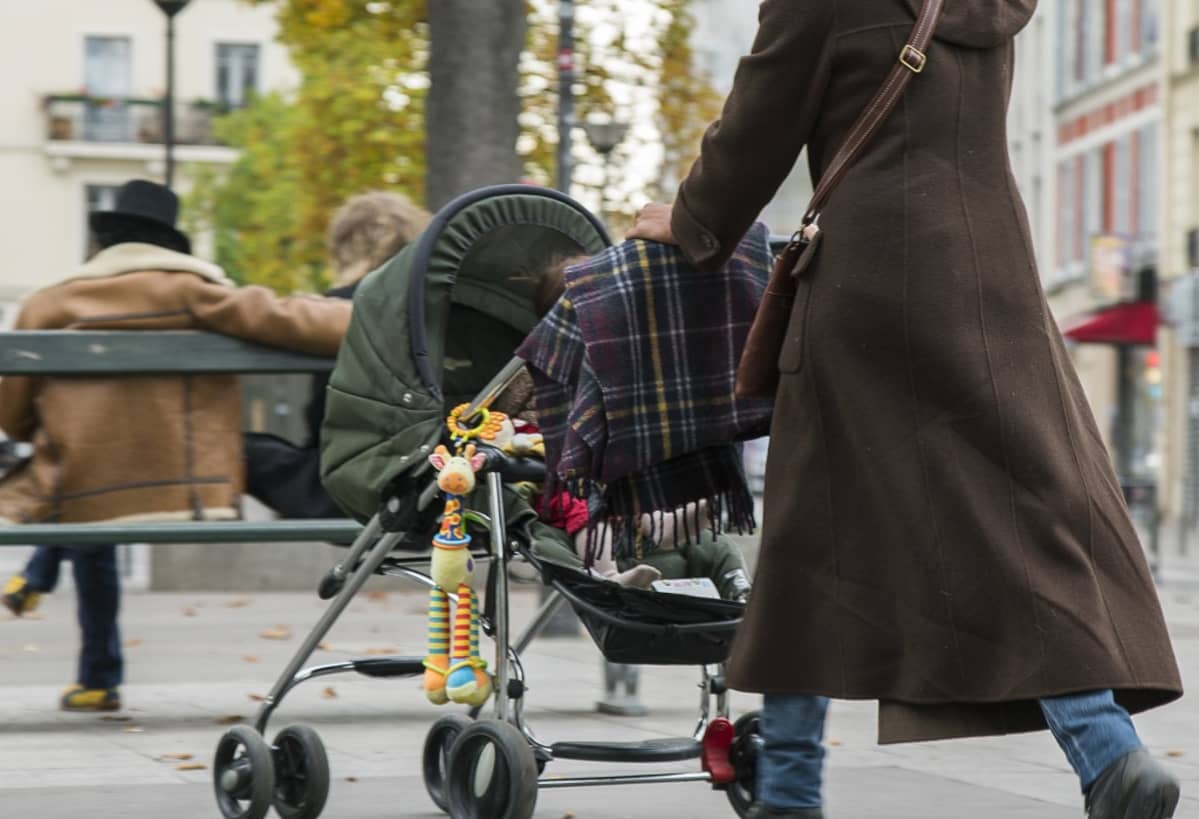 Äiti lykkää vauvaa vaunuissa, joista roikkuu värikäs lelu.