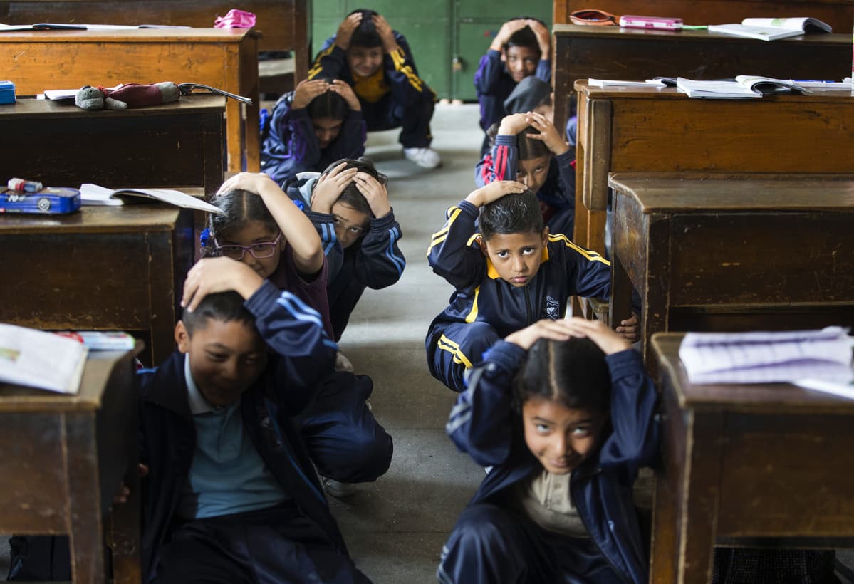 Nepalilaiset koululaiset istuvat kyykyssä koululuokassa ja suojelevat käsillään päätään maanjäristysharjoituksen aikana.