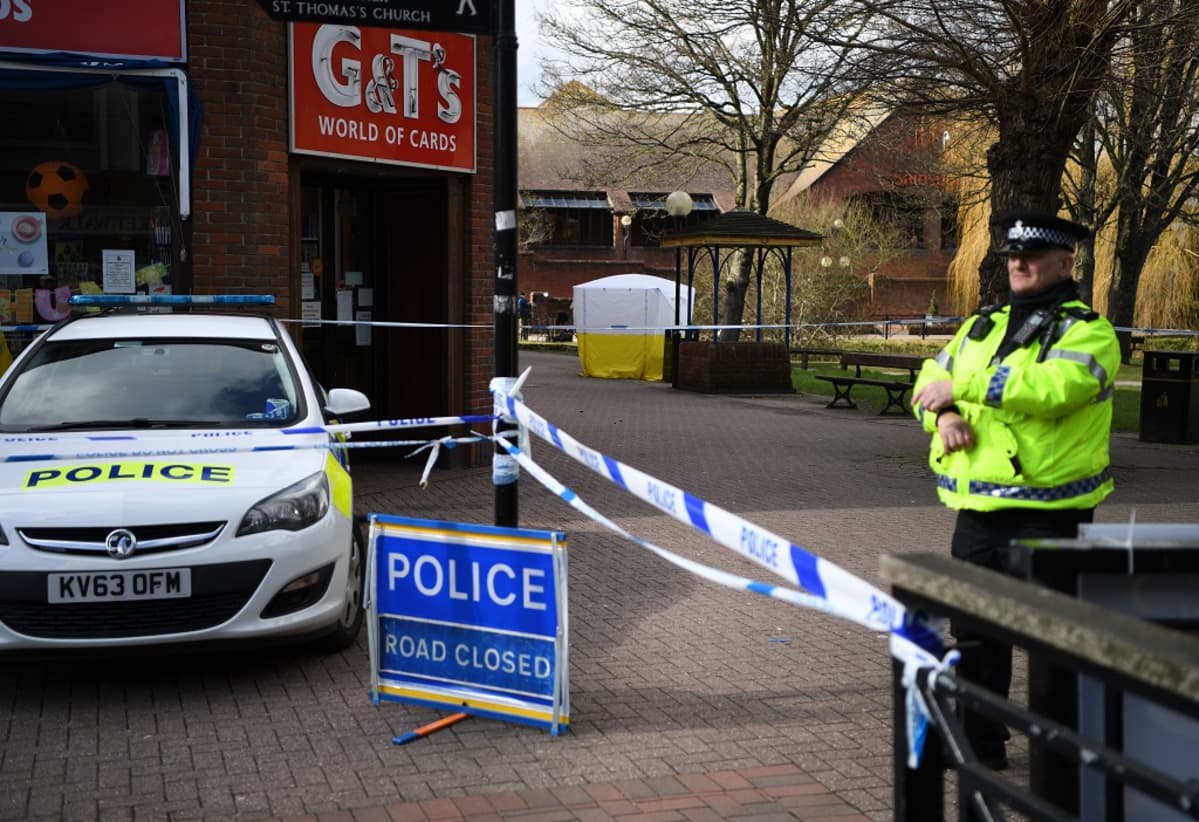 Keltaiseen huomioliiviin pukeutunut brittipoliisi seisoo vartiossa poliisin nauhalla eristetyn alueen laidalla kuvan oikealla reunalla. Kuvan vasemmalla puolella on poliisiauto parkkeerattuna. Auton vieressä on maassa kyltti, jossa lukee valkoisin kirjaimin sinisellä pohjalla: "Poliisi. Tie suljettu." Taustalla näkyy valko-keltainen teltta, jolla on peitetty yksi puiston penkeistä.