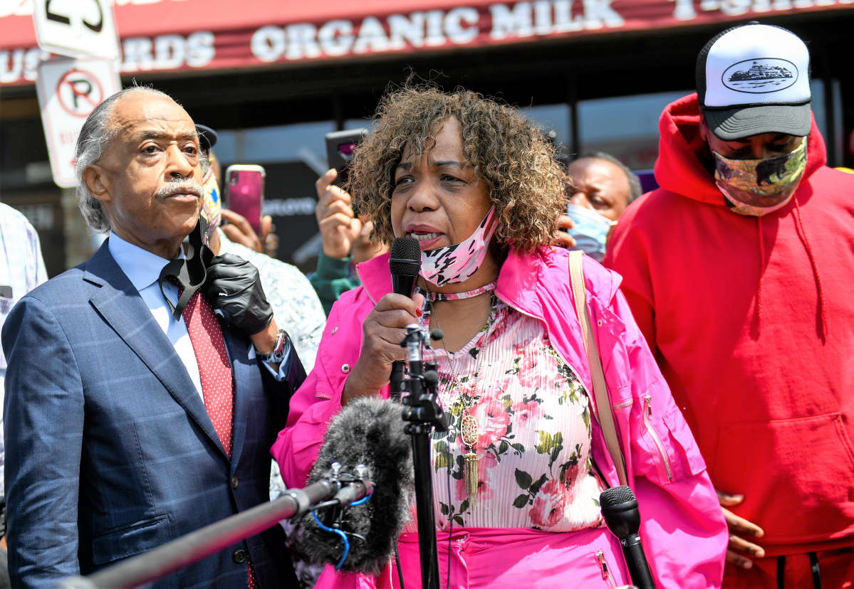 Eric Garnerin äiti Gwen Carr puhuu mikrofoniin. Al Sharpton kuuntelee vakavana vieressä. Taustalla näkyy väkijoukkoa ja elintarvikekaupan julkisivua.