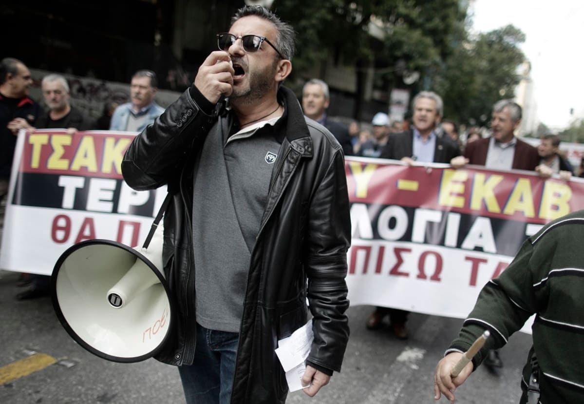 Mielenosoitusta. Etualalla mustaan nahkatakkiin pukeutunut parrakas mies mustissa silmälaseissa puhuu megafonin mikrofoniin. Taustalla joukko mielenosoittajia kantaa plakaattia, jossa on kreikankielistä kirjoitusta.