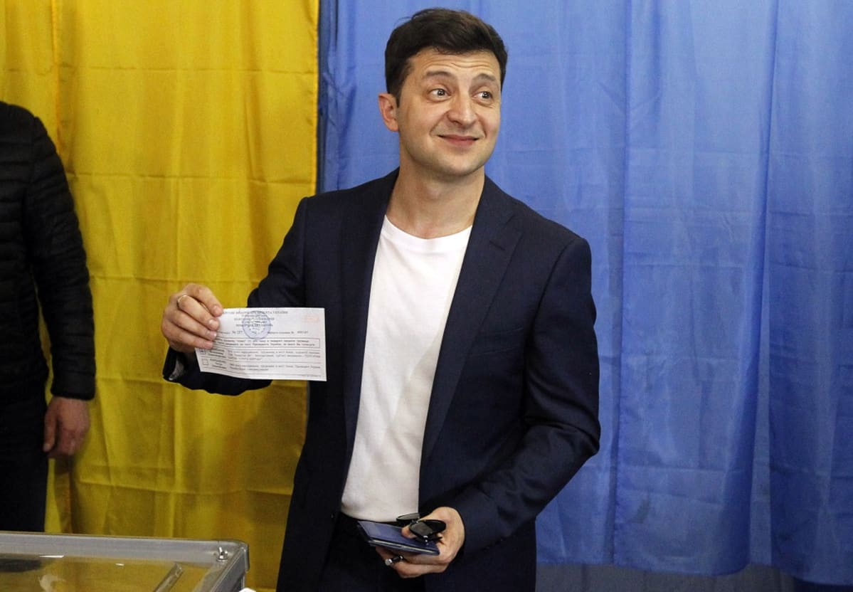 Hymyilevä Zelenskyi näyttää äänestyslipukettaan toimittajille. Taustalla on Ukrainan lipun värinen kangas.