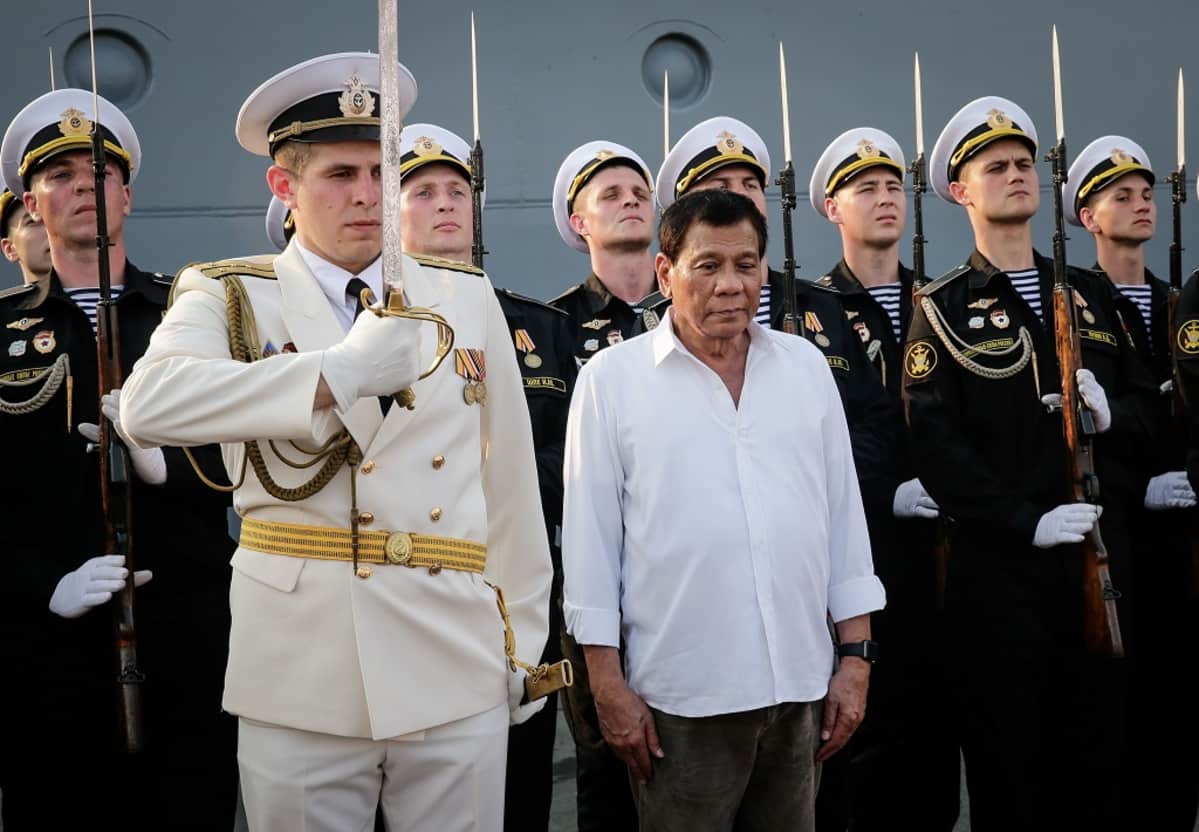 Presidentti Duterte seisoo valkoisessa kauluspaidassa paraatiunivormuissa olevien venäläisten laivastosotilaiden rivin edessä. Hänen vieressään seisoo upseeri valkoisessa univormussa ja koppalakissa pitäen miekka koholla. Taustalla olevilla merisotilailla on valkoiset koppalakit, mustat univormut ja kiväärit, joissa on pistimet kiinnitettyinä.