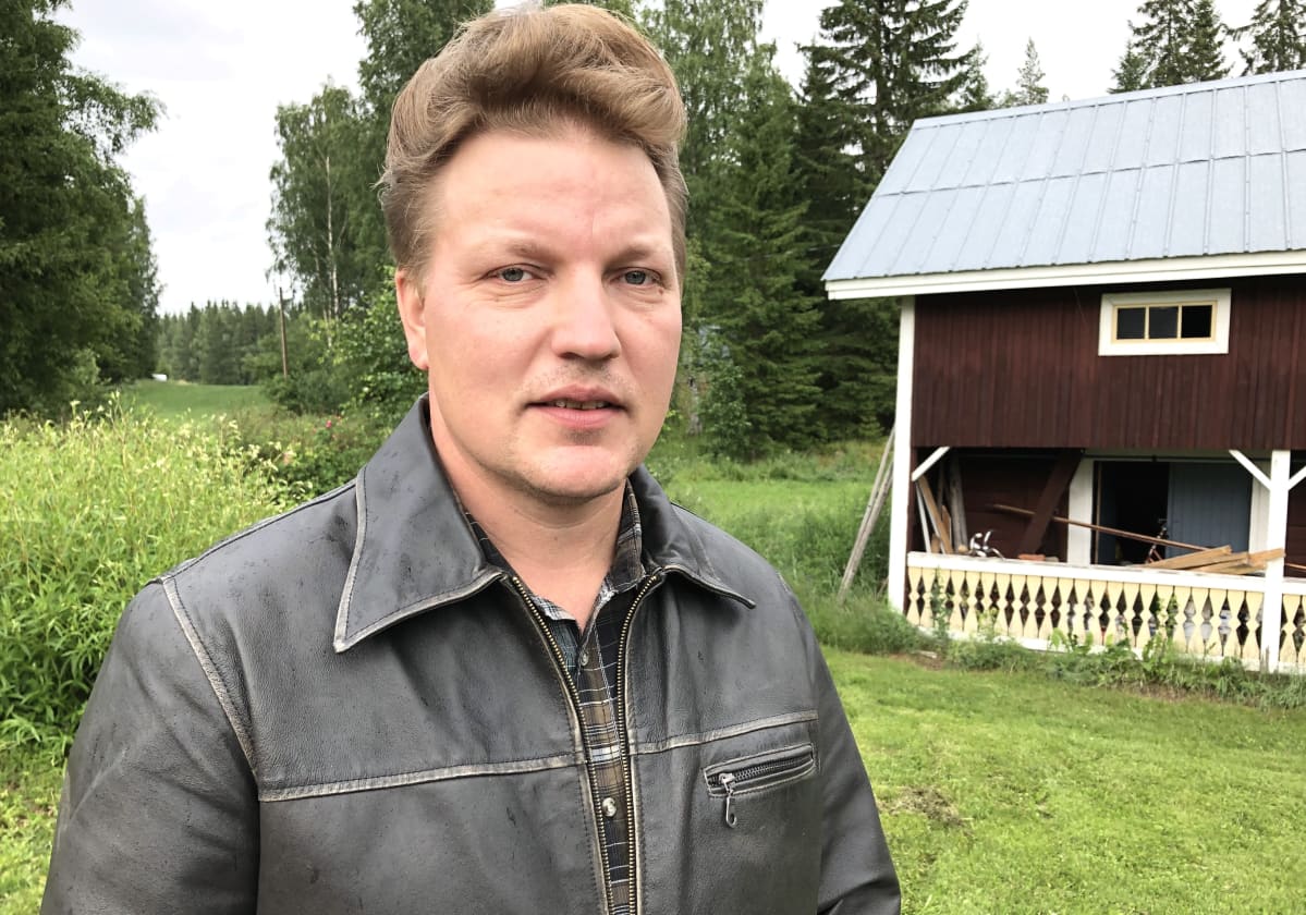 Mies seisoo piha-aitan edessä, nahkatakki päällä, ja katsoo kameraan.