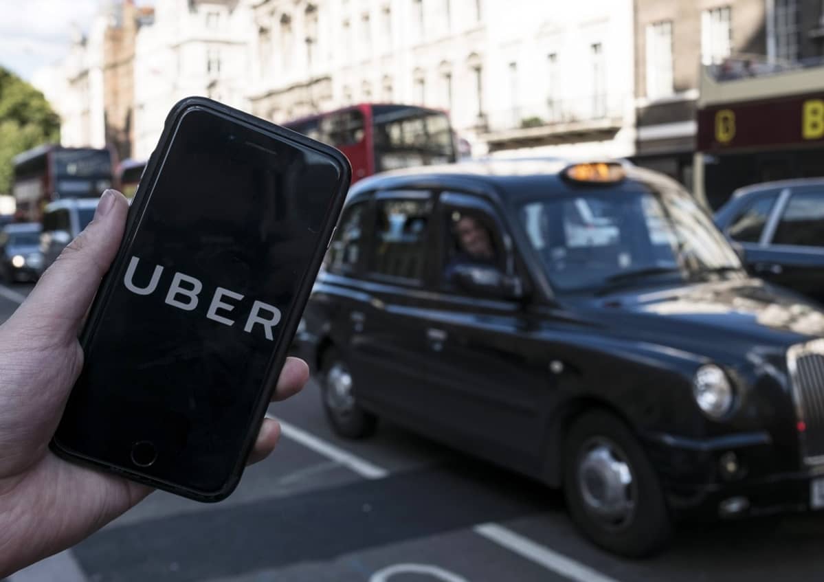 Etualalla näkyy älykännykkä, jonka näytöllä lukee isoilla kirjaimilla UBER. Taustalla näkyy musta Lontoon taksi.