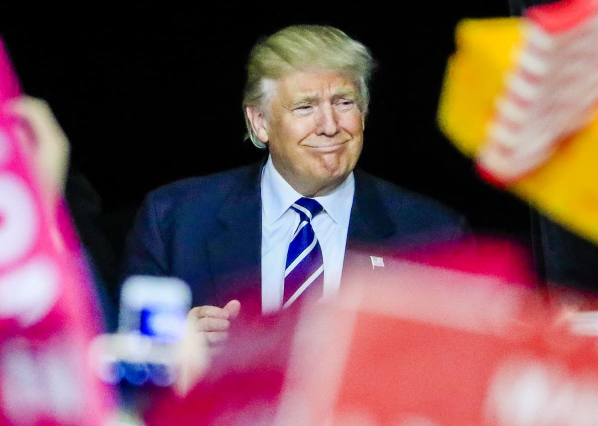Trump hymyilee kuvan keskellä sinisessä puvussaan ja sinivalkoisessa solmiossa.