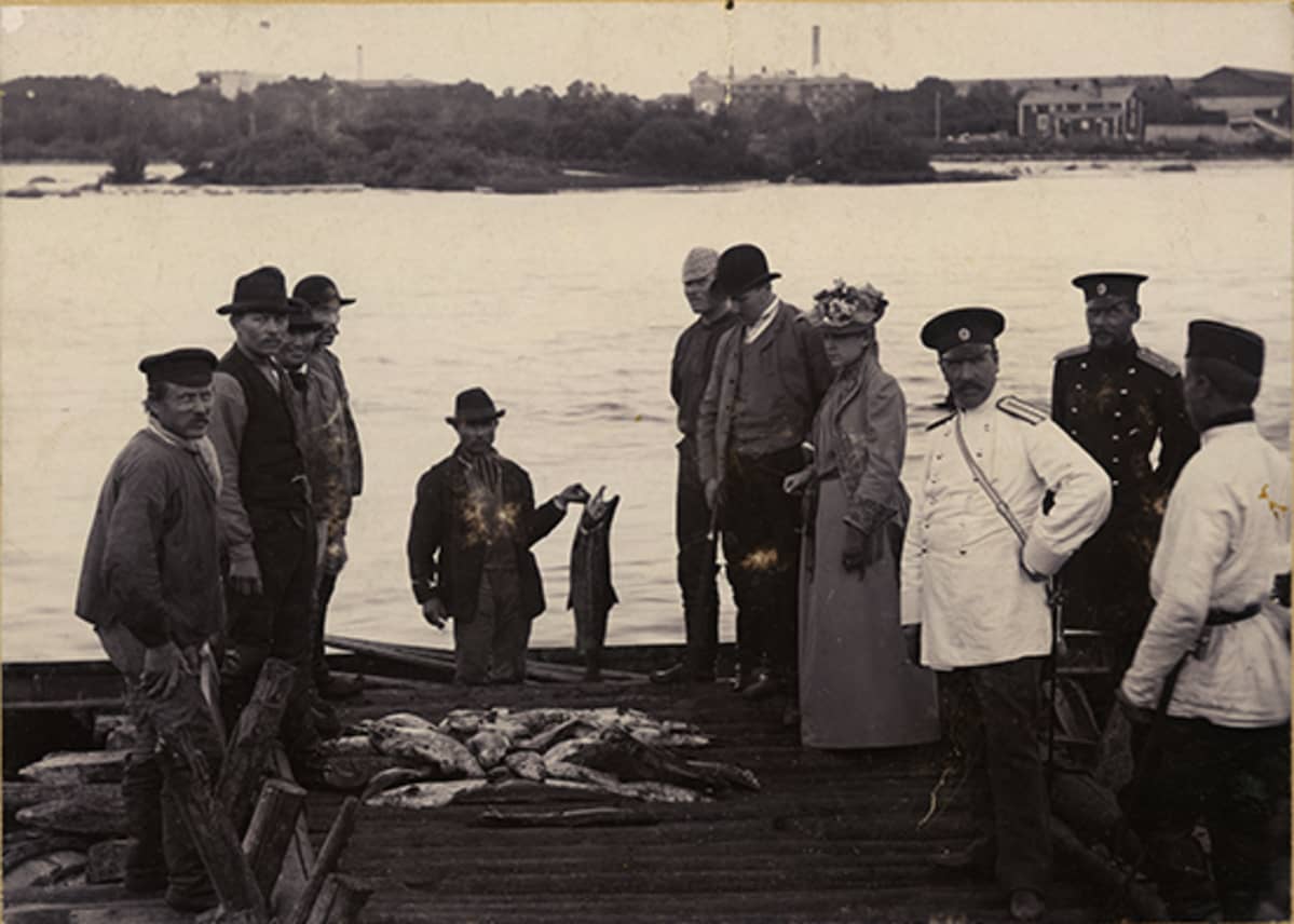 Mustavalkoisesa valokuvassa ihmisiä Oulujoen rannalla ihailemassa lohisaalista.