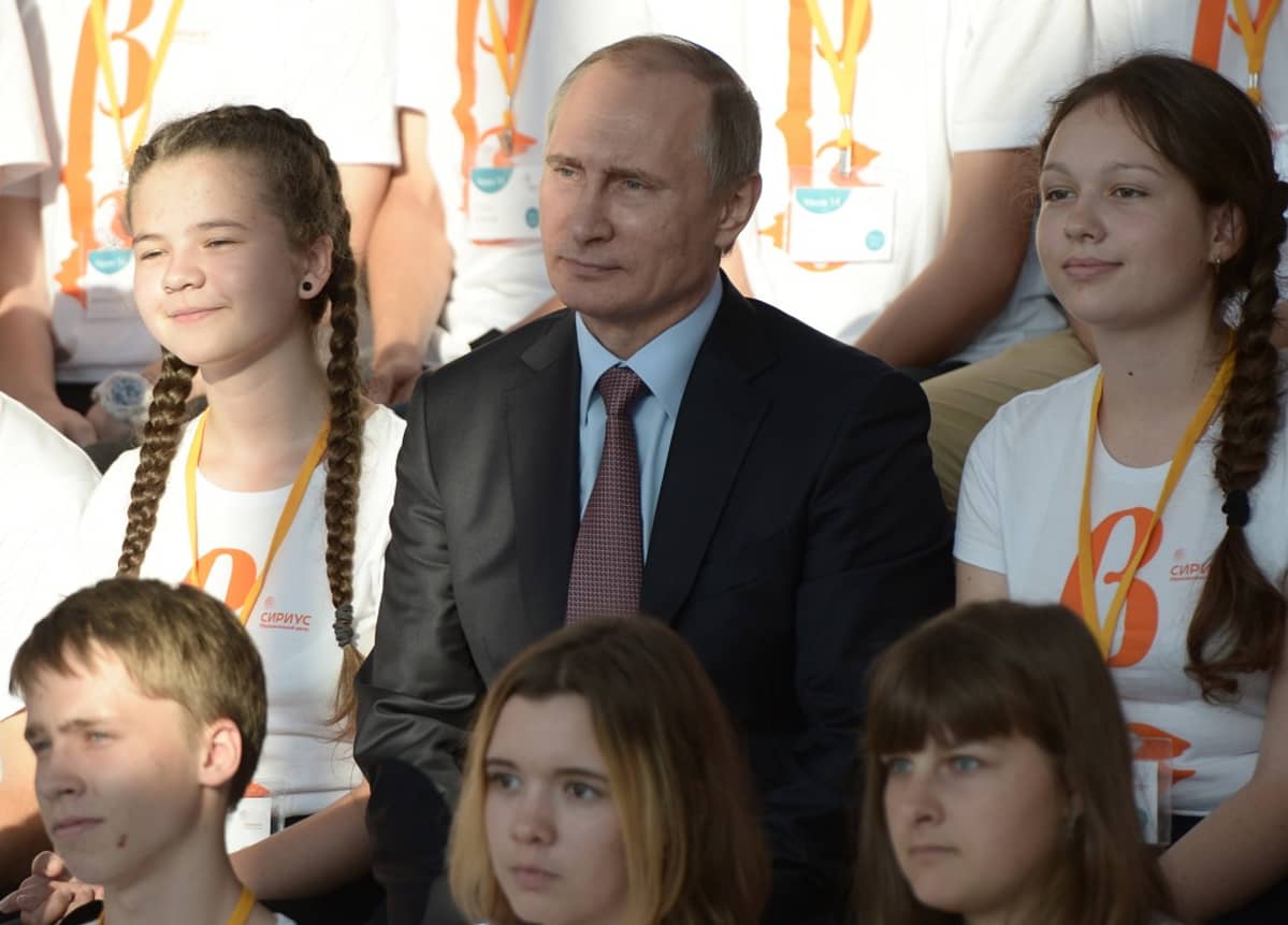 Vladimir Putin istumassa kahden lettipäisen tytön välissä.