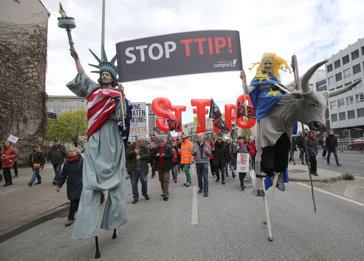 Vapaudenpatsaaksi pukeutunut mies ja EU:ta symboloiva nainen kantavat kylttiä, jossa lukee "Stopp TTIP!" Taustalla muita mielenosoittajia lippuineen.