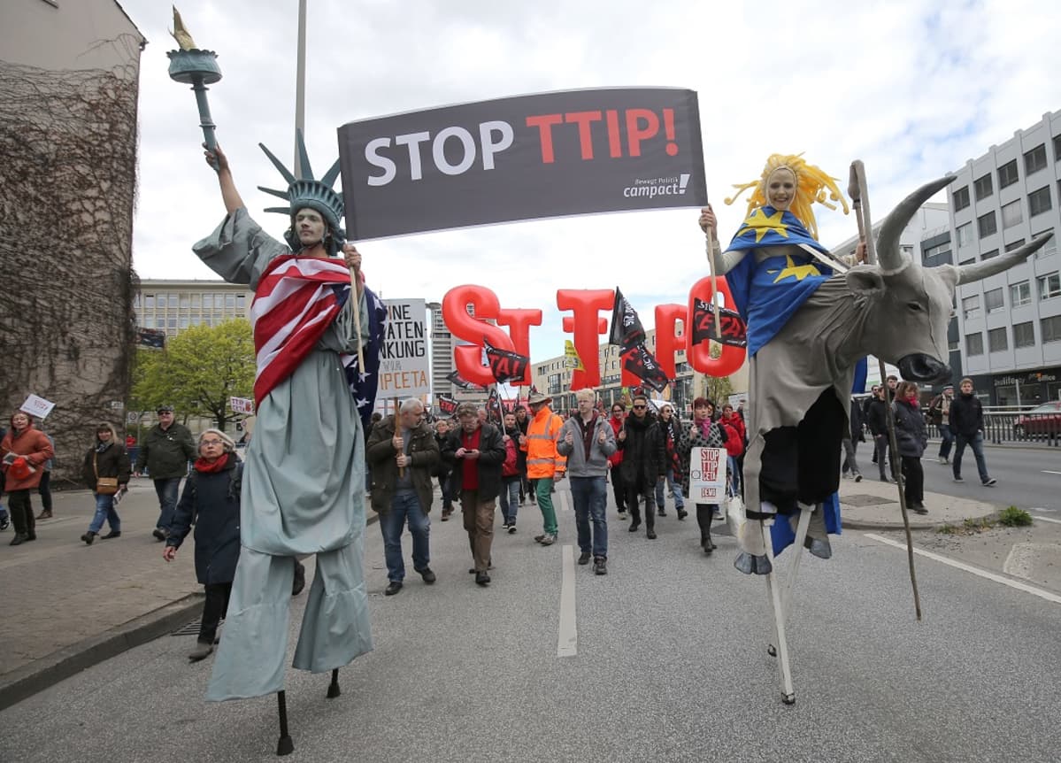 Vapaudenpatsaaksi pukeutunut mies ja EU:ta symboloiva nainen kantavat kylttiä, jossa lukee "Stopp TTIP!" Taustalla muita mielenosoittajia lippuineen.