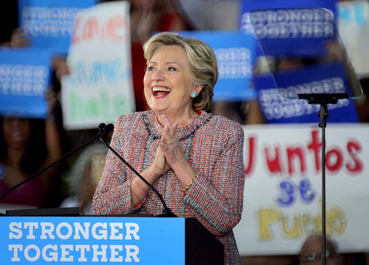 Hillary Clinton puhujakorokkeen takana. Taustalla ihmiset pitelevät häntä tukevia banderolleja.