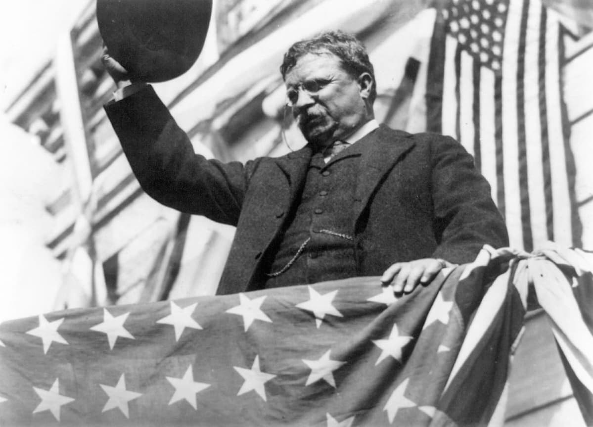 Presidentti Theodore Roosevelt tervehtii yleisöä Yhdysvaltain lipun peittämällä korokkeella.