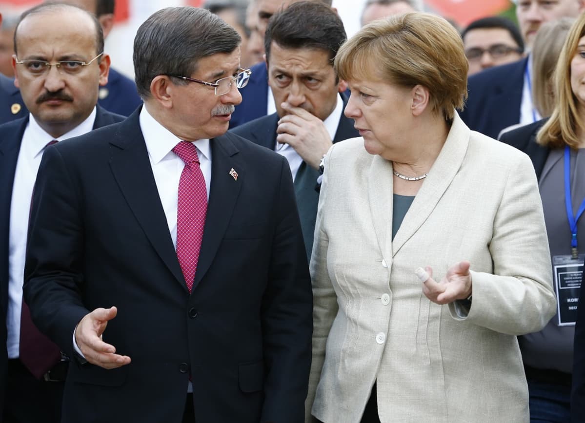 Ahmet Davutoğlu ja Angela Merkel keskustelevat. Turkin pääministerillä on tumma puku ja punainen ruudukas kravatti, Saksan liittokanslerilla vaalea bleiseri, vihreä paita. Taustalla näkyy virkamiehiä.