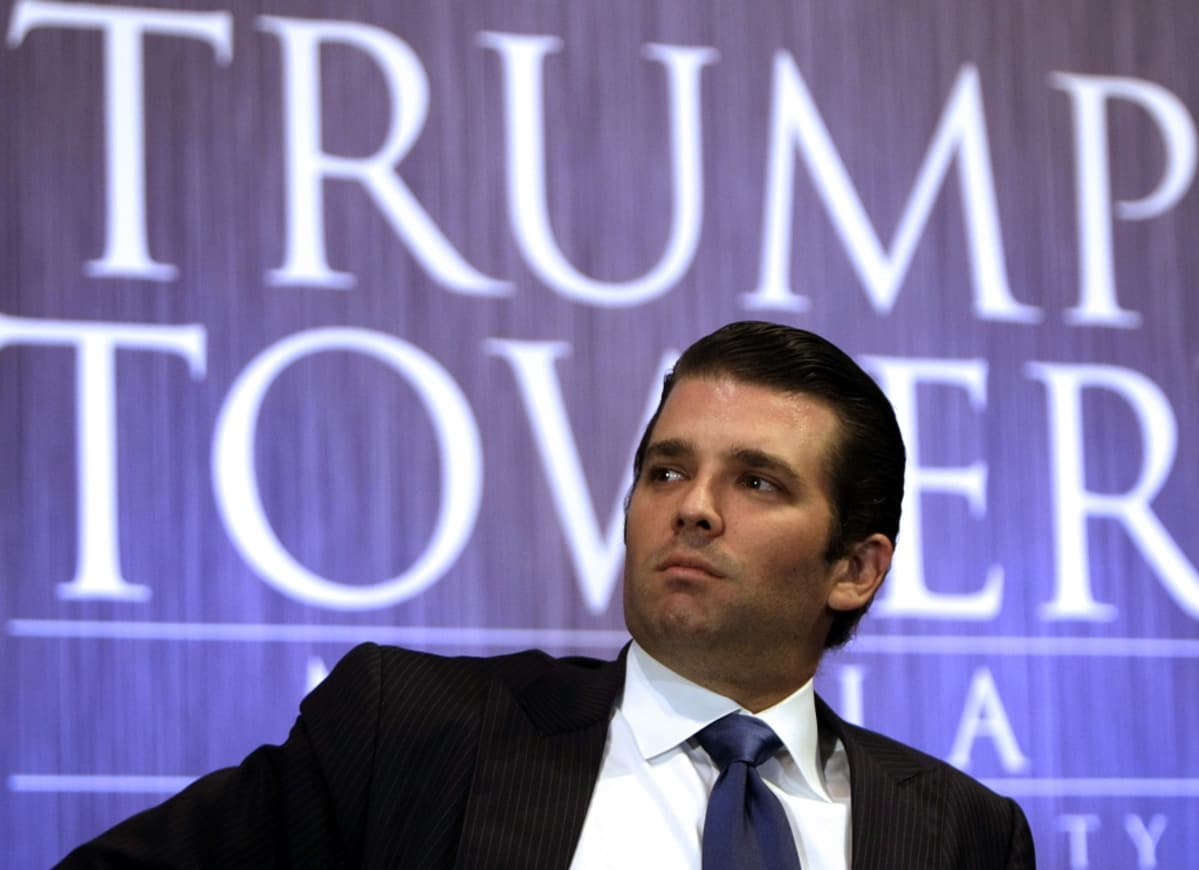 Trump on tummassa puvussa, tumma kravatti kaulussaan istuvassa asennossa. Taustalla näkyy iso teksti Trump Tower sinisellä pohjalla.