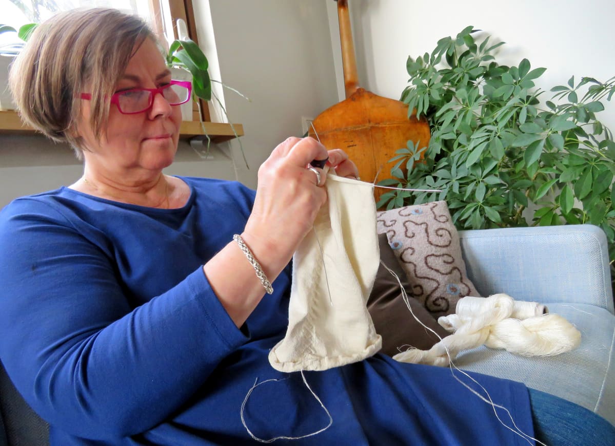 Liisa Kylmänen istuu sohvalla neulomassa silkkisukkaa ohuenohuilla puikoilla,taustalla huonekasveja ja nurkassa sello.