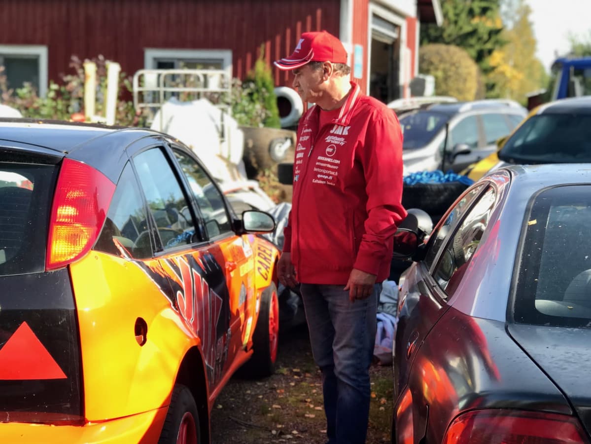 Punaiseen takkiin ja lippikseen pukeutunut rallikuski mies seisoo ralliautojen keskellä