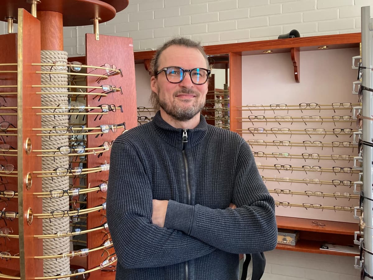 Silmälasipäinen hymyilevä mies seisoo kkädet puuskassa optikkoliikkeessä.