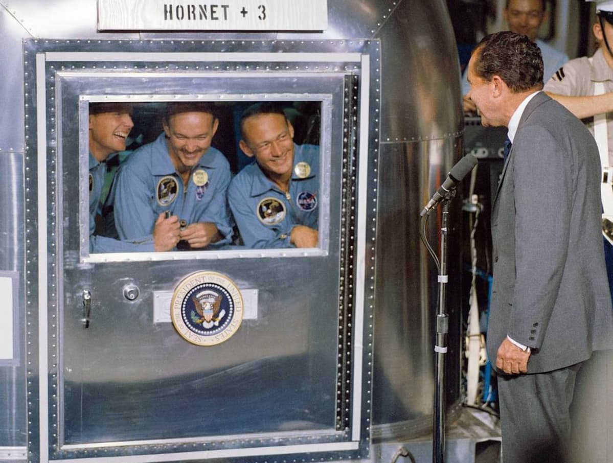 Arnstrong, Collins ja Aldrin kurkistevat hymyillen karanteenihuoneen ikkunasta, jonka edessä seisoo Nixon.