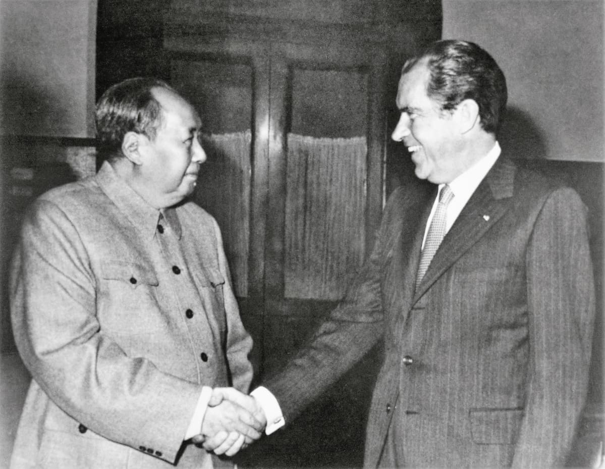 Mao Zeodong ja Richard Nixon kättelevät hymyillen. Maolla on univormunkaltainen yksinkertainen takki, Nixonilla puku, jossa on liituraitoja ja kravatti.Kuva on mustavalkoinen.